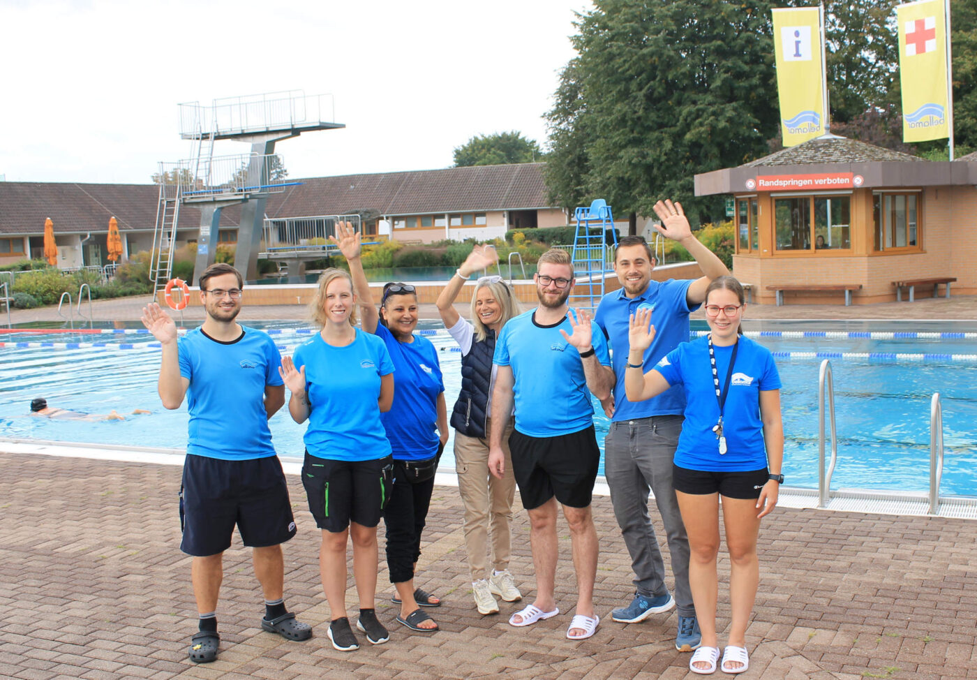 Das Bellamar-Team steht lachend und winkend vor dem Schwimmbecken mit dem Sprungturm im Hintergrund